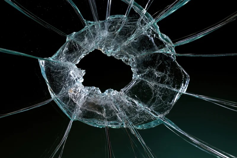 Shattered glass pane with central impact hole and radiating cracks against dark background