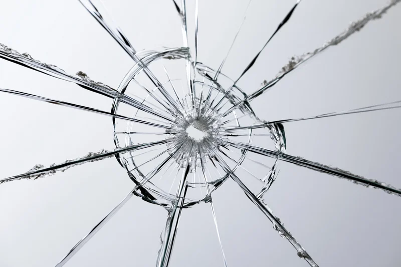 Shattered glass pane with radial cracks from central impact point against plain background