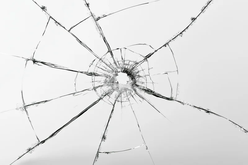 Shattered glass pane with central impact hole radiating cracks across transparent surface