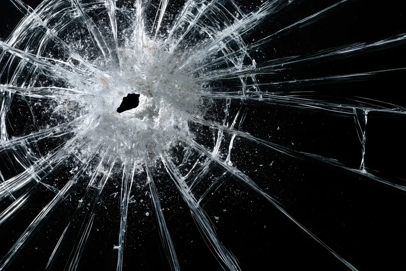 Shattered glass pane with central impact hole and radiating cracks against black background