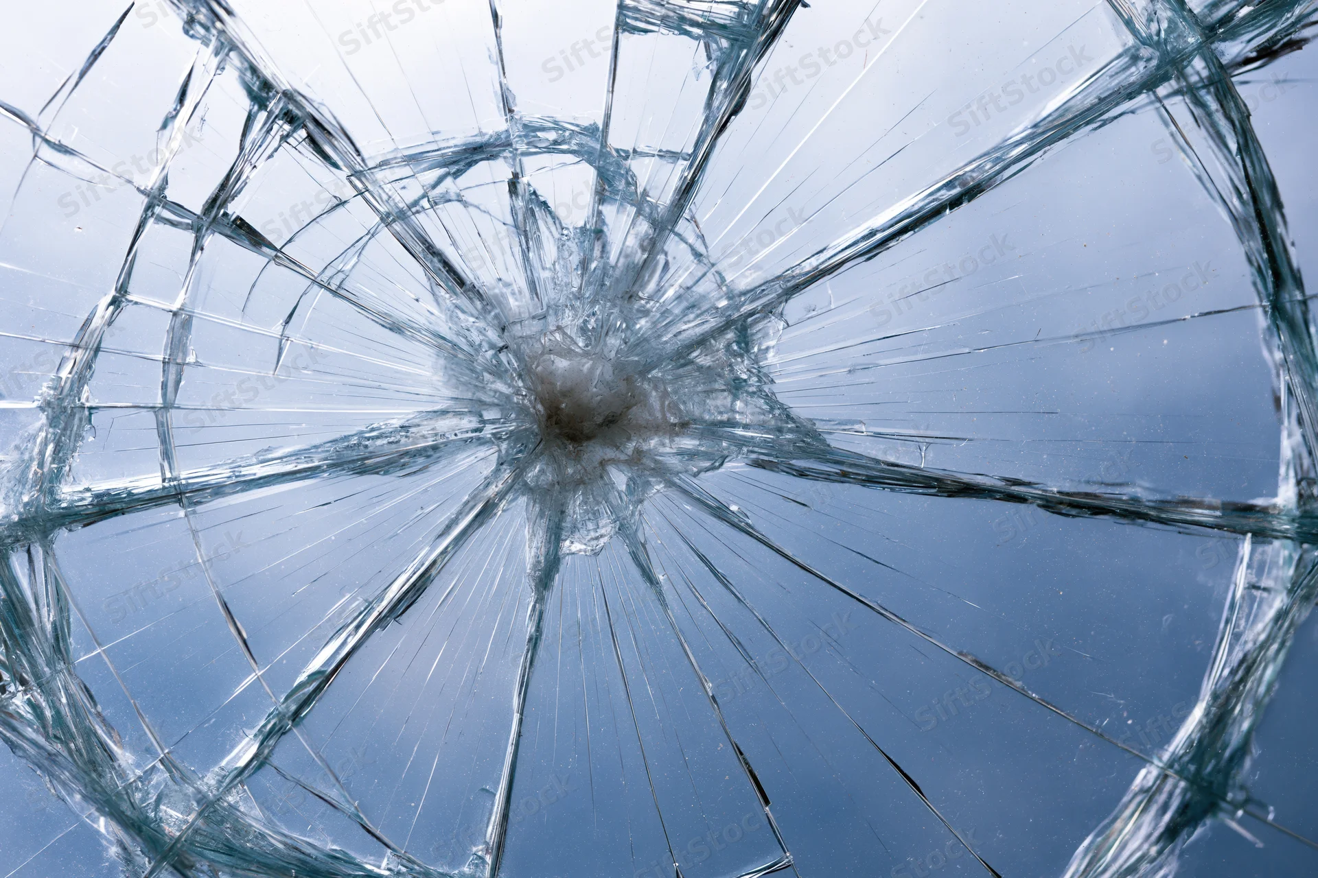 Shattered glass pane with radial cracks from central impact point against blue sky