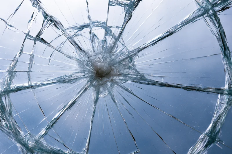 Shattered glass pane with radial cracks from central impact point against blue sky