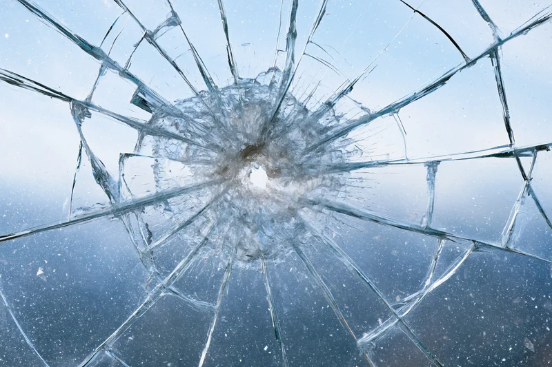 Shattered glass pane with radial cracks from central impact point against blue sky background