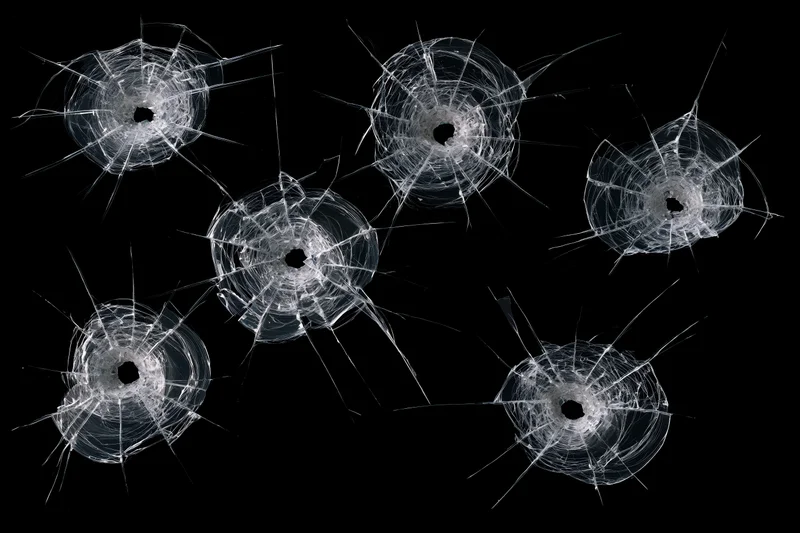 Six bullet holes in a dark tempered glass pane each with radial cracks and a central impact point