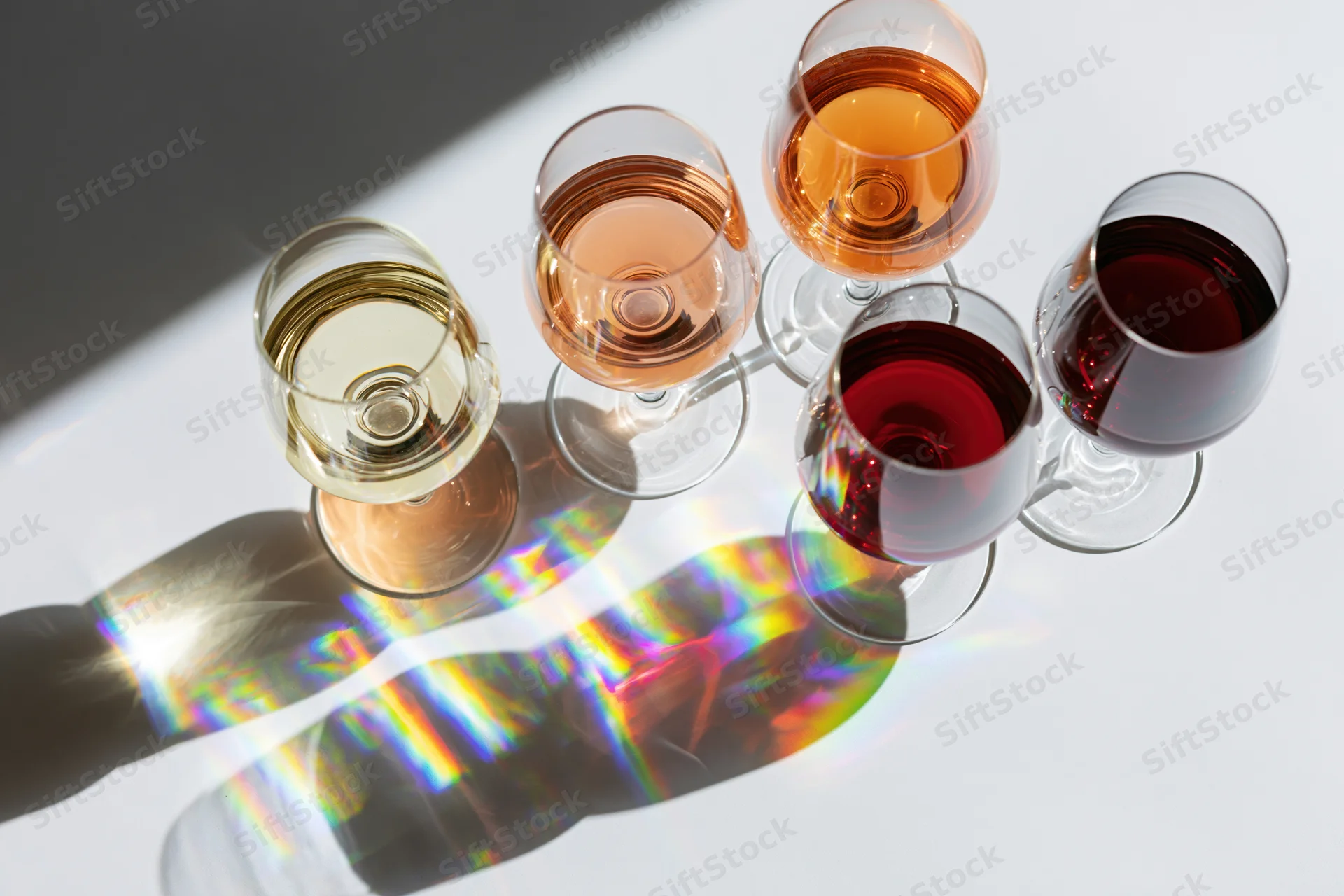 Six wine tasting glasses arranged diagonally on a white surface with rainbow light reflections