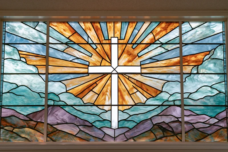 Stained glass window with white christian cross radiating light over mountains and clouds in church interior