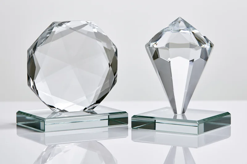 Two blank crystal glass trophies side by side on square bases with faceted octagonal and diamond shapes, reflecting light on a white surface