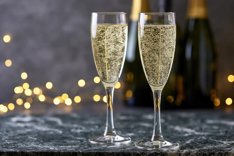 Two champagne flutes filled with sparkling wine and rising bubbles on a dark marble surface with blurred festive lights behind