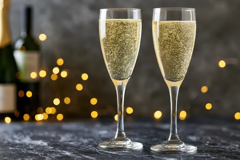 Two champagne flutes filled with sparkling wine and rising bubbles on a dark stone table with blurred festive lights in the background