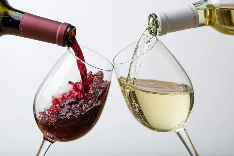 Red and white wine being poured simultaneously into tilted stemmed glasses against a plain white background