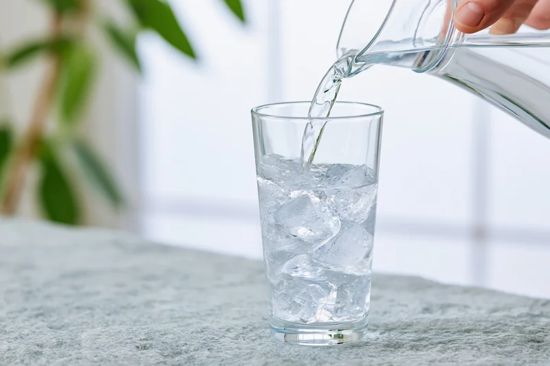 Water being poured from a glass pitcher into a tall glass with ice on a countertop