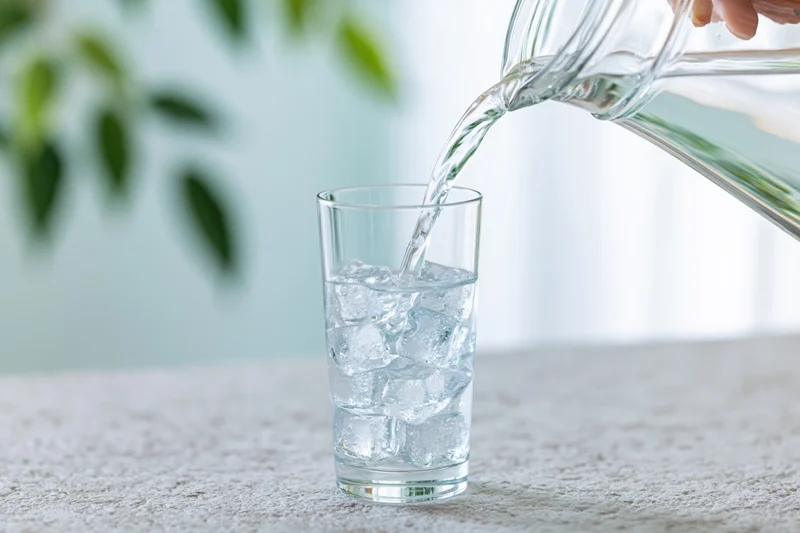 Water being poured from a glass pitcher into a tall glass with ice on a tabletop