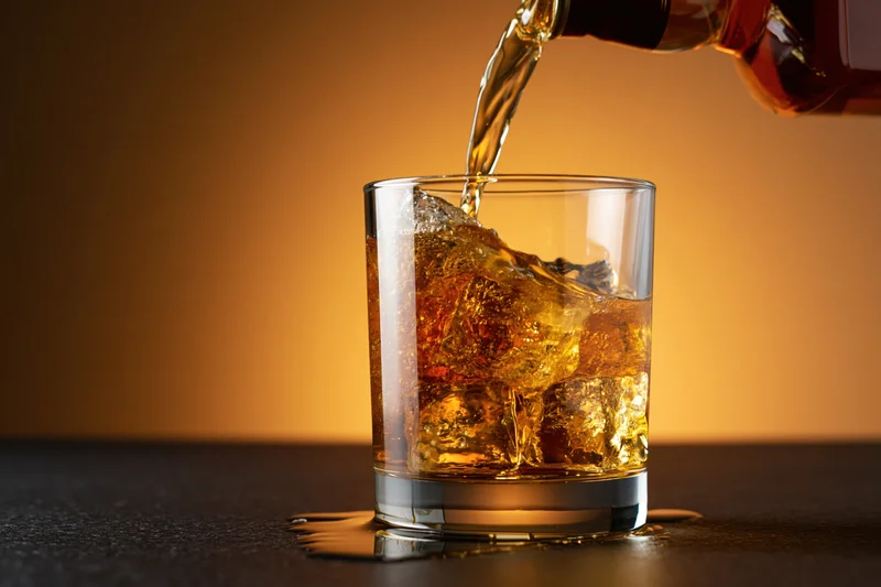 Whiskey being poured into a rocks glass with ice on a dark surface