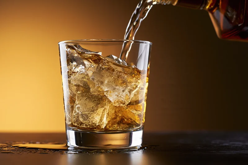 Whiskey being poured into a rocks glass filled with ice on a dark surface