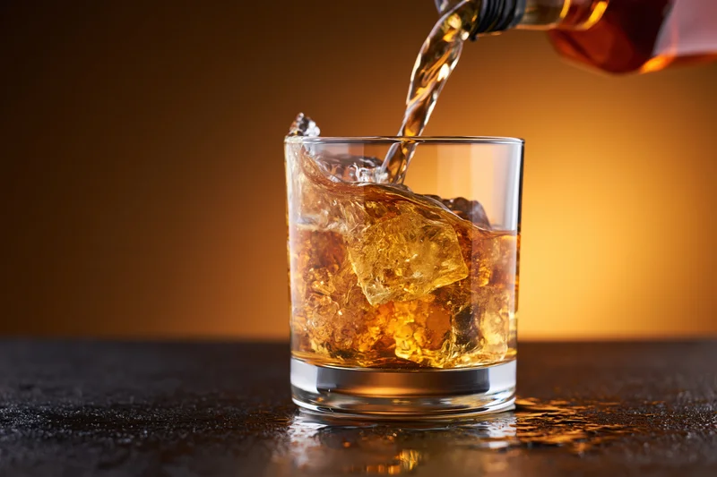 Whiskey being poured into a rocks glass filled with ice on a dark surface