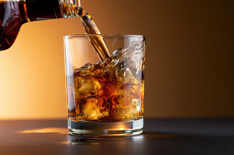 Whiskey being poured into a rocks glass with ice on a dark surface