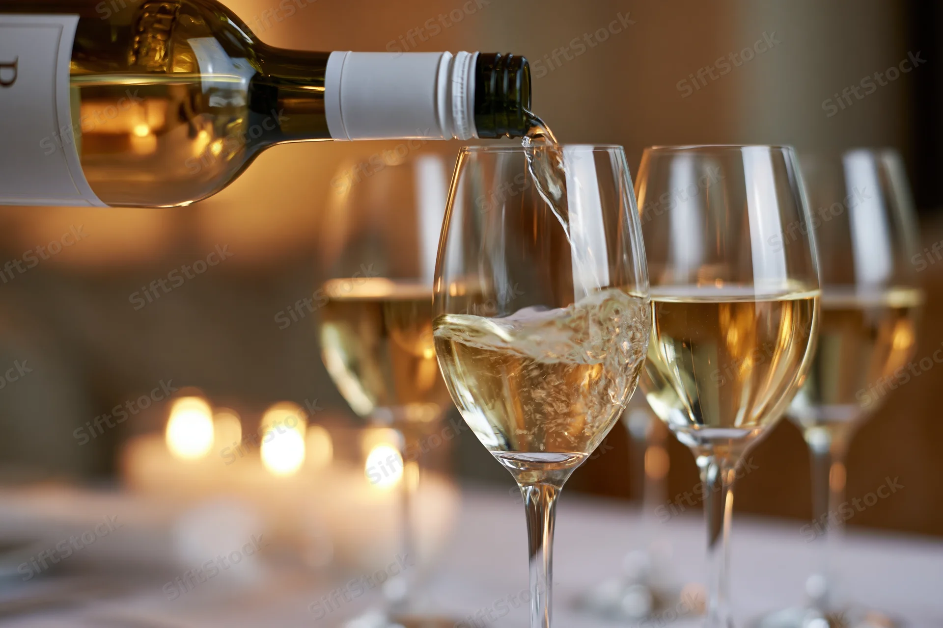 White wine being poured from a green glass bottle into a crystal wine glass at a dinner table with candlelight in the background
