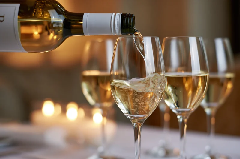 White wine being poured from a green glass bottle into a crystal wine glass at a dinner table with candlelight in the background
