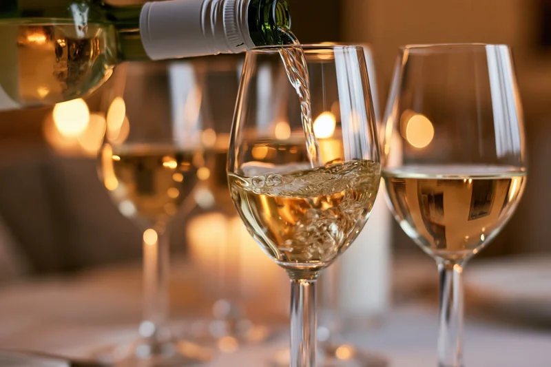 White wine being poured from a green bottle into a crystal glass at a dinner table with warm ambient lighting