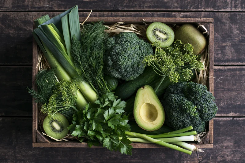 Wooden crate filled with assorted green vegetables and fruits arranged on straw on dark wood surface