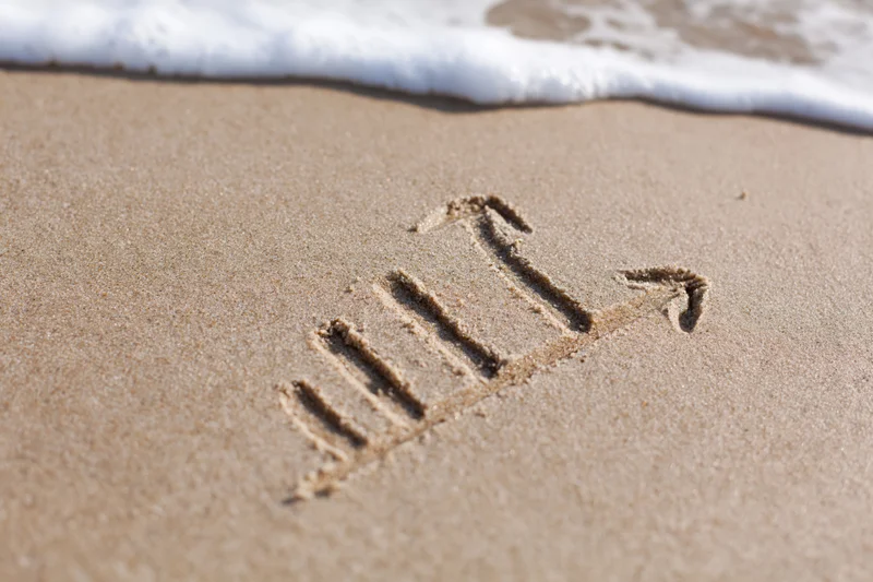 Ascending bar chart with arrows drawn in sand near ocean wave