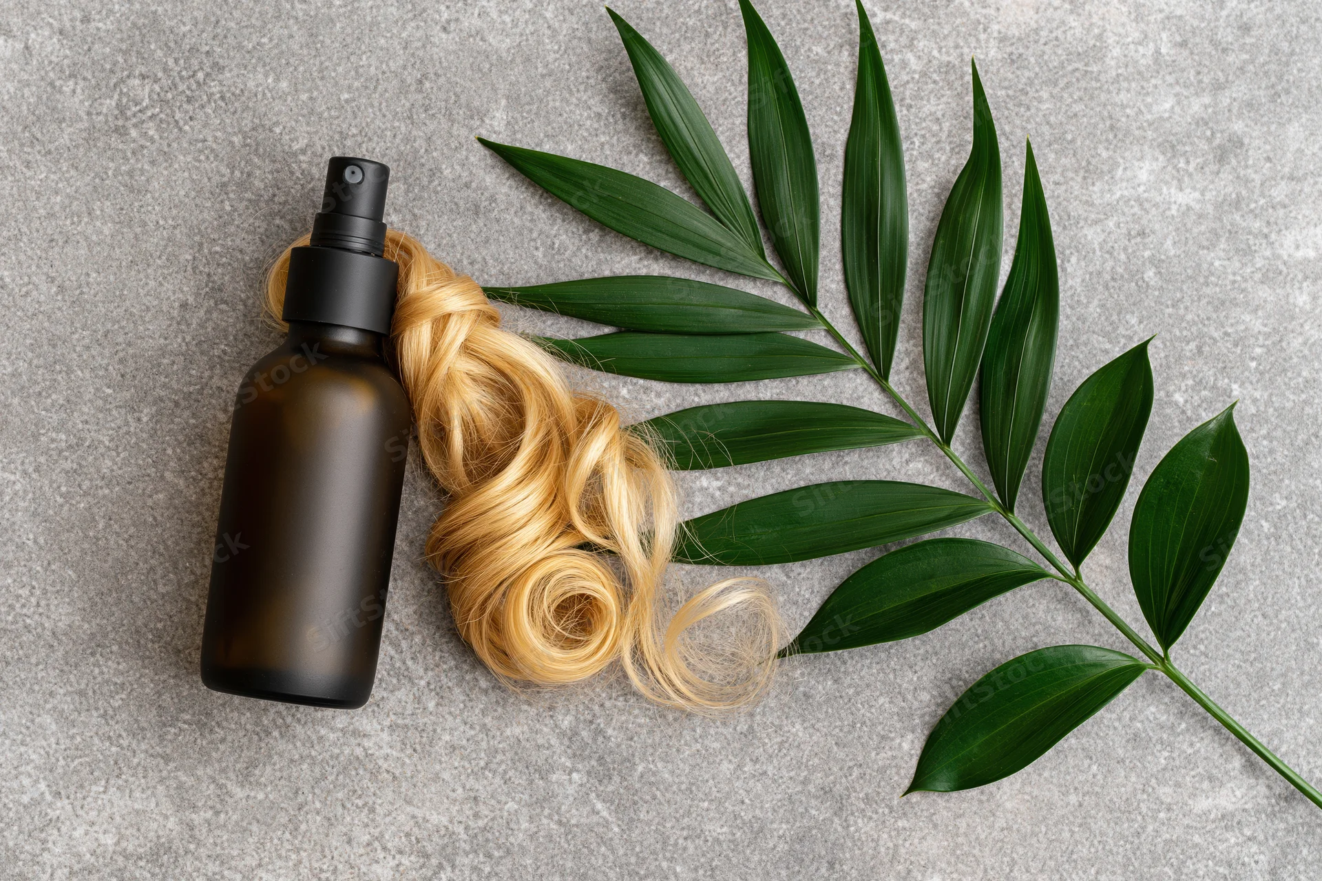 Dark amber spray bottle for hair care lying flat on gray surface beside blonde curly hair and green leaf