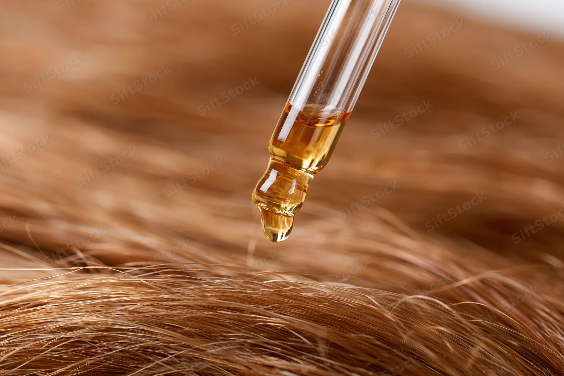 Glass dropper releasing golden oil onto blonde hair strands in closeup view