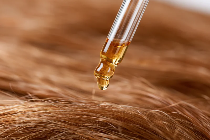 Glass dropper releasing golden oil onto blonde hair strands in closeup view