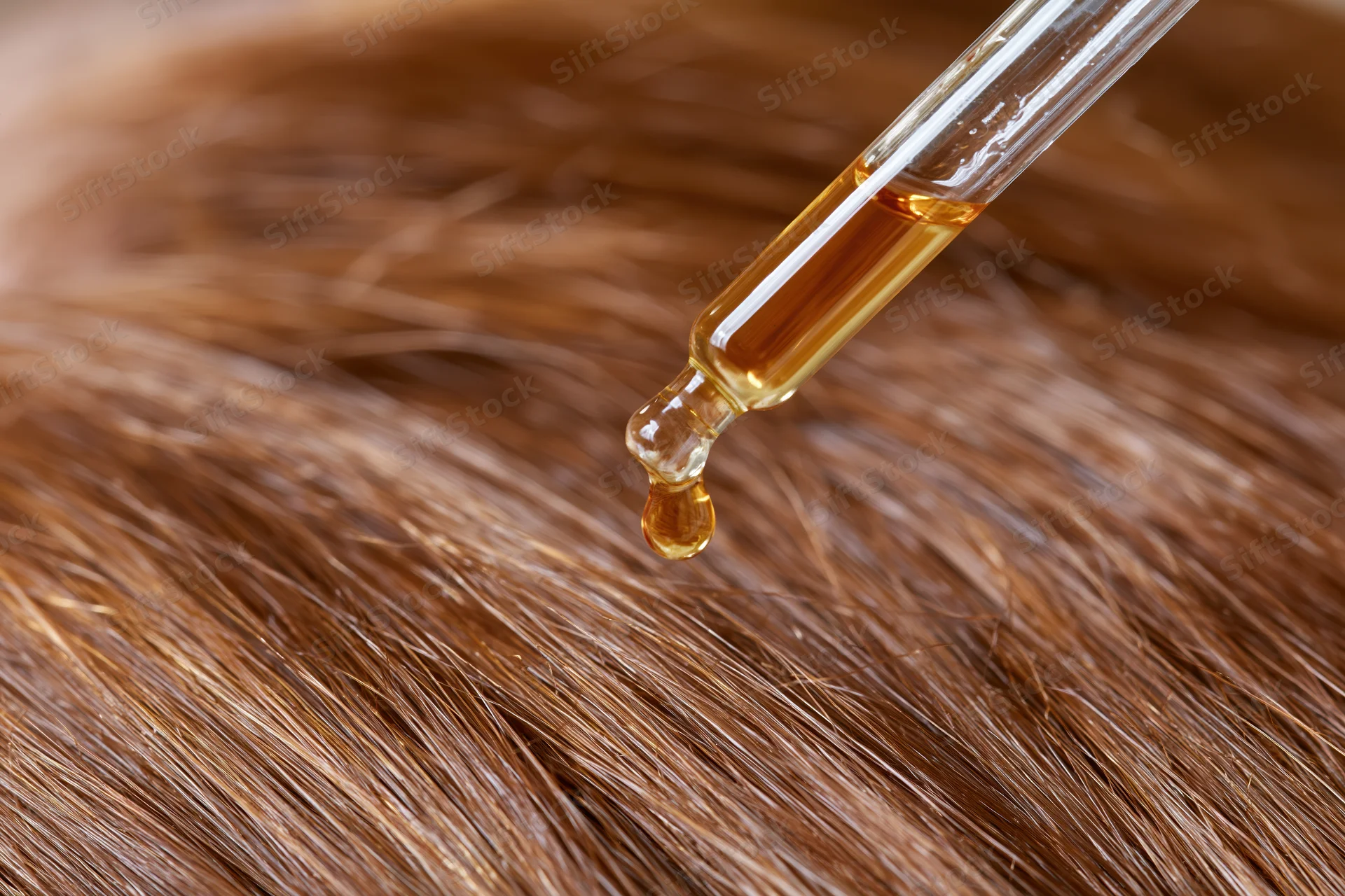 Golden oil drop from glass dropper falling onto brown hair strands