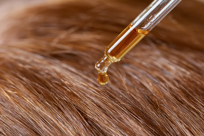Golden oil drop from glass dropper falling onto brown hair strands