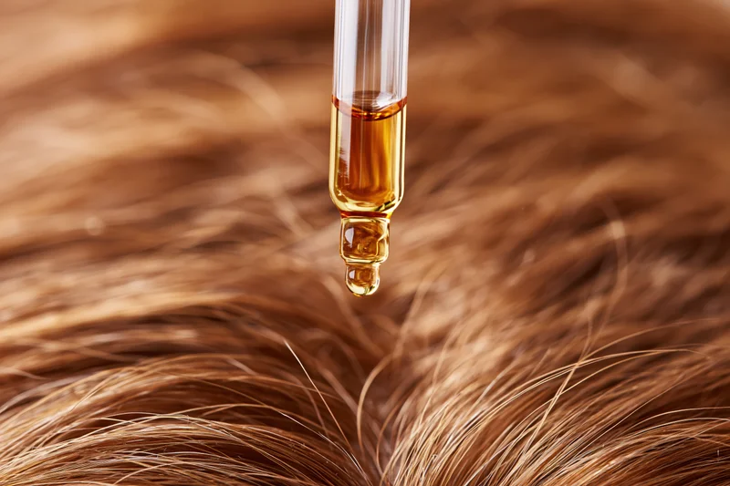 Golden oil drop falling from glass dropper onto brown hair strands