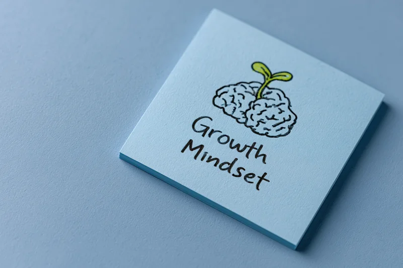 Light blue sticky note with hand-drawn brain and seedling labeled growth mindset on flat blue surface