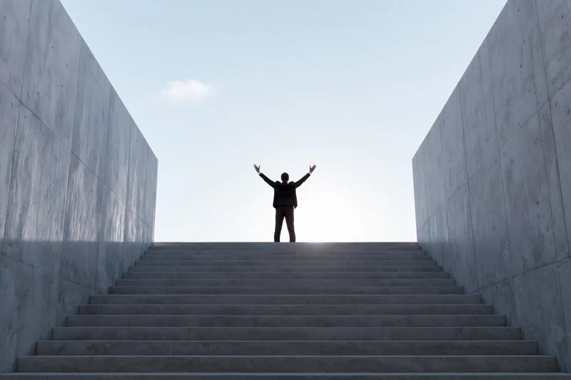 Man in silhouette with arms raised at top of concrete staircase under bright sky