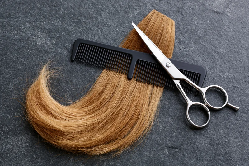 Honey-blonde hair curl with scissors and comb on dark grey slate surface