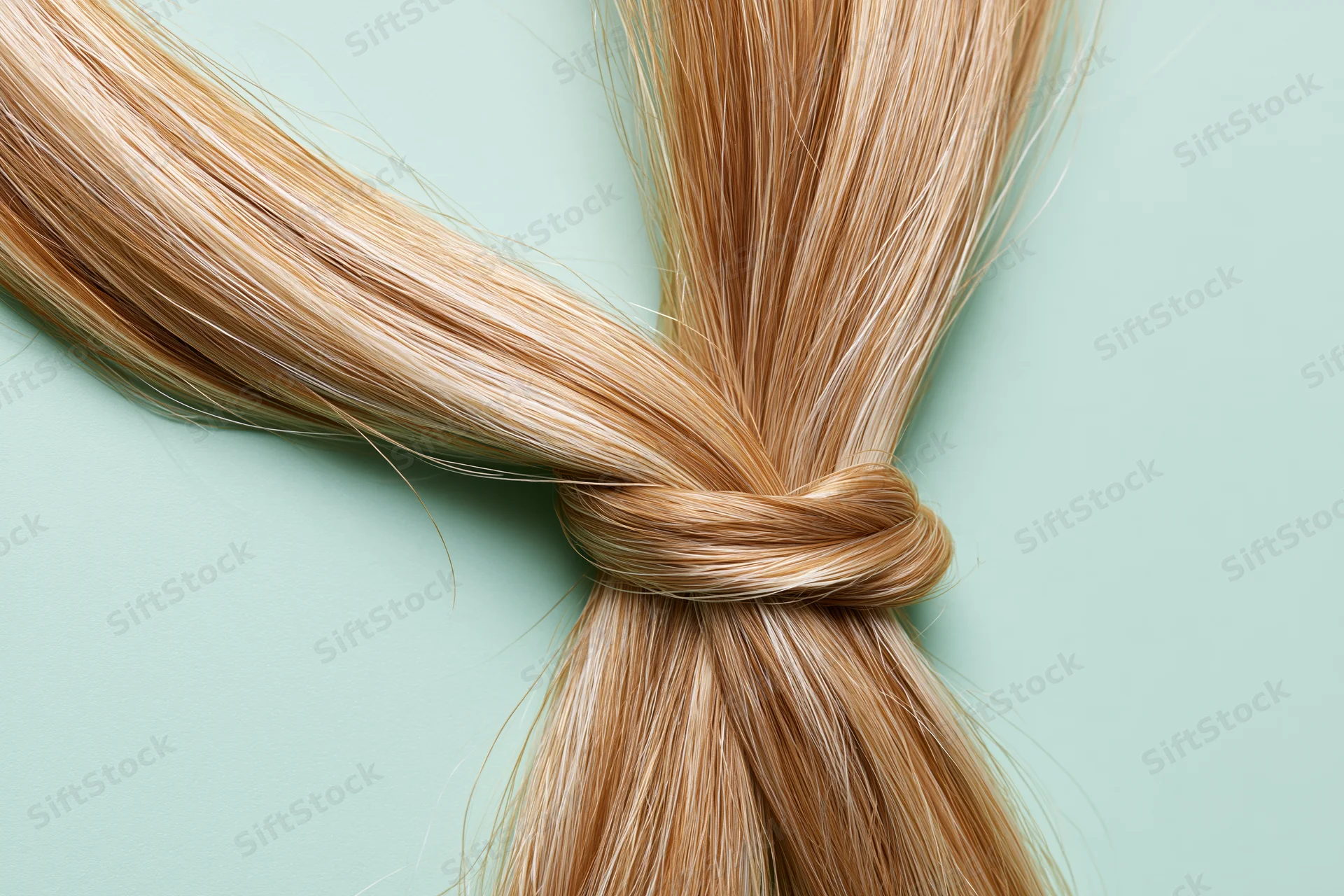 Thick blonde hair strand tied in a loose knot laid flat on a light green surface
