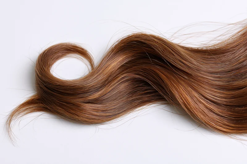 A loose wavy strand of medium brown hair with warm auburn highlights lying on a white surface