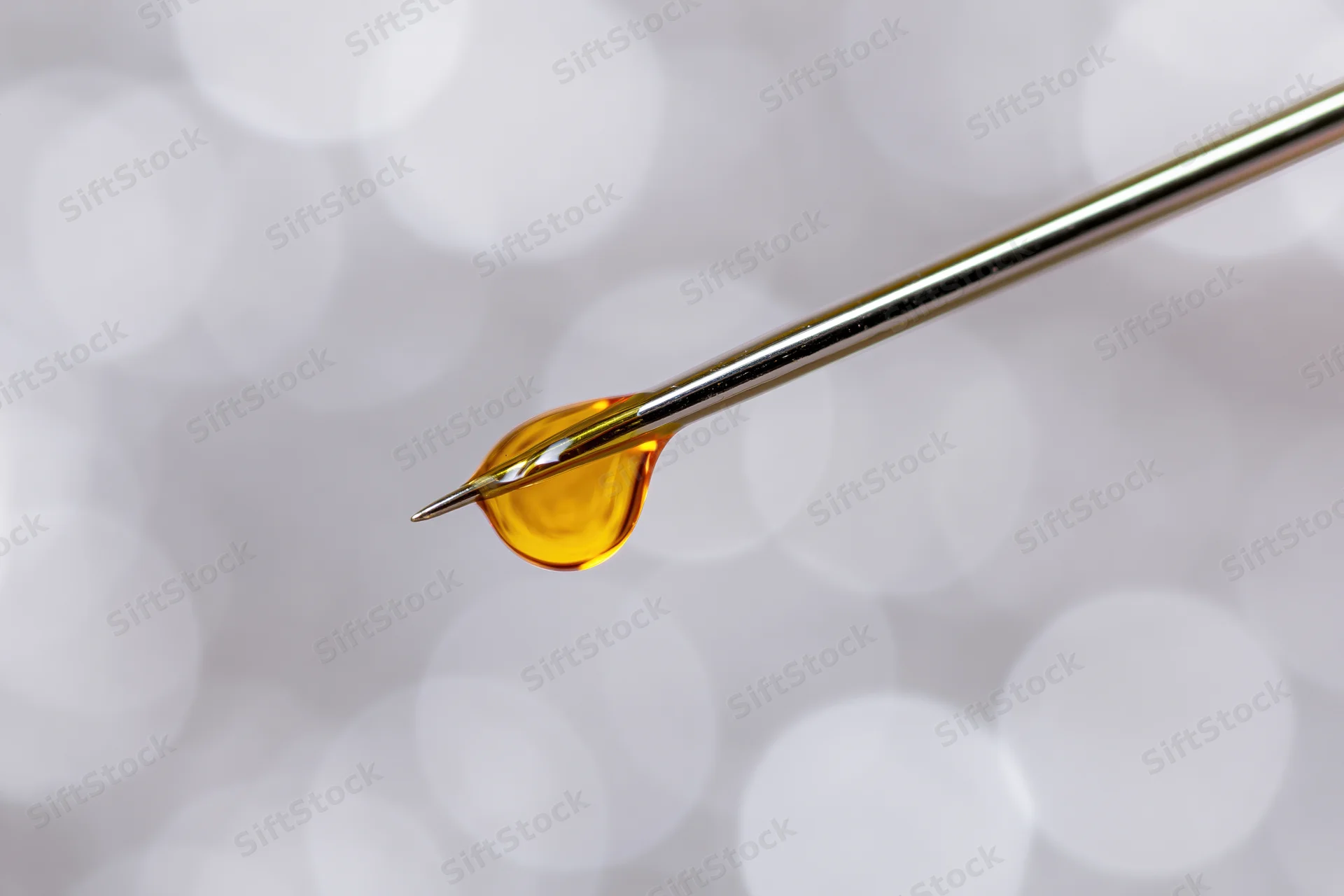 Golden oil droplet suspended on sharp needle tip against soft white bokeh background