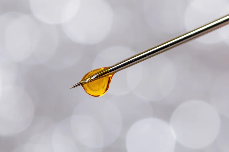 Golden oil droplet suspended on sharp needle tip against soft white bokeh background