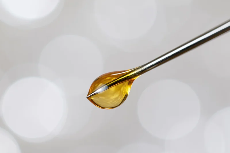 Golden oil droplet suspended on a syringe needle tip against a soft bokeh background