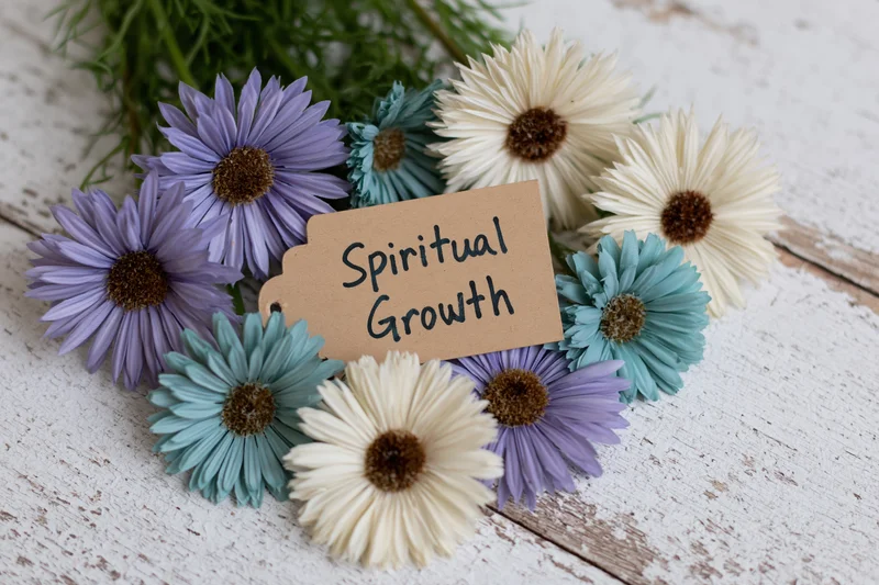 Kraft paper tag with handwritten spiritual growth text surrounded by purple blue and cream daisies on white wooden surface