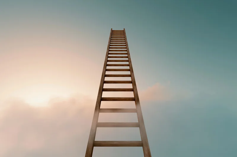 Wooden ladder rising vertically into the sky above soft clouds at dawn