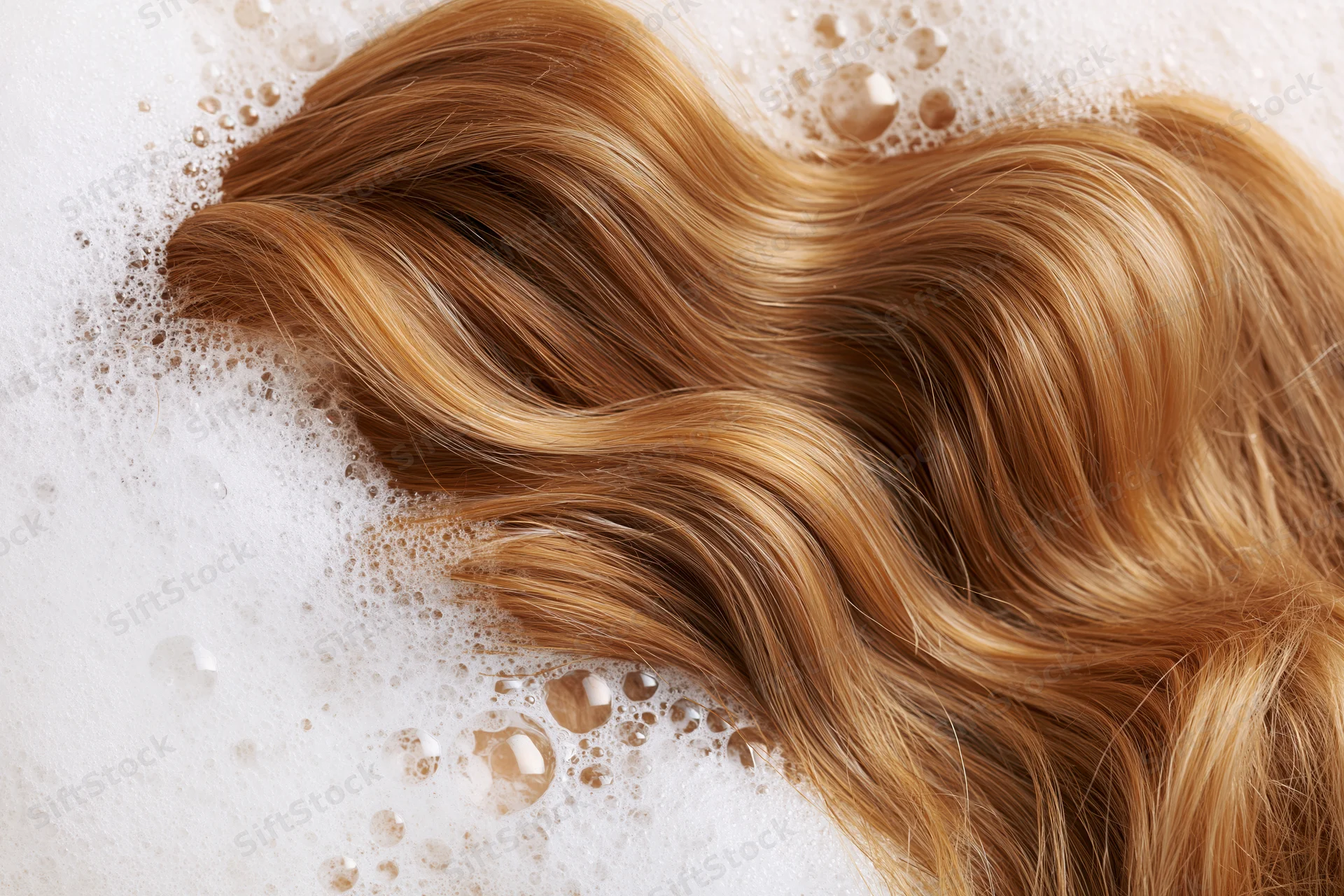 Wavy golden-brown hair strand surrounded by shampoo foam