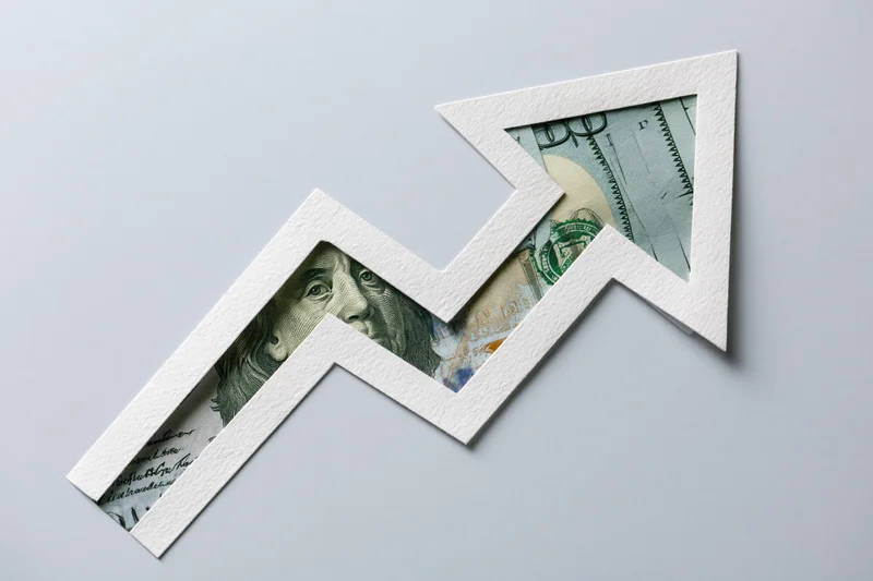 White paper arrow cutout revealing dollar bills inside, forming an upward zigzag trend on a light gray surface