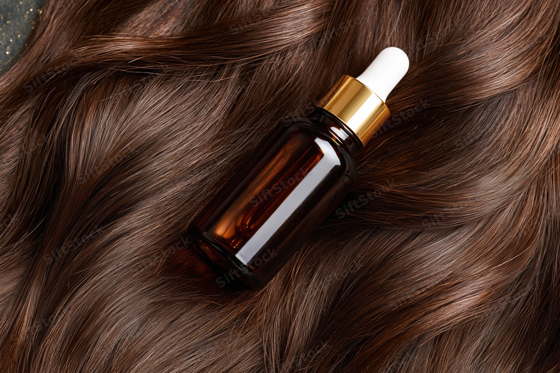 Amber glass dropper bottle with gold collar and white rubber cap resting on long brown hair