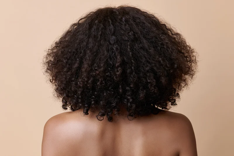 Back of a black woman with voluminous curly hair and bare shoulders against a neutral background
