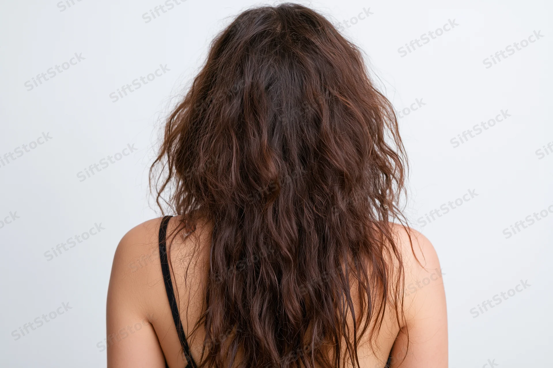 Back view of a young woman with long frizzy wavy dark brown hair wearing a black spaghetti strap top against a white background