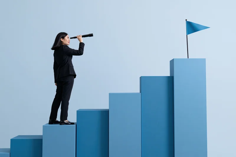 Businesswoman in black suit using telescope while standing on blue steps toward a flag at the top