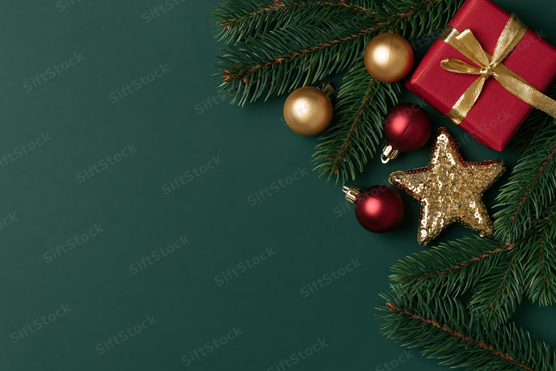 Christmas decorations including pine branches, red gift box, gold and red ornaments, and a sequined star arranged on a dark green surface with copyspace