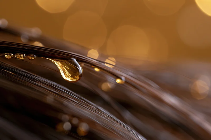 Close-up of oil droplets on dark hair strands with warm bokeh background