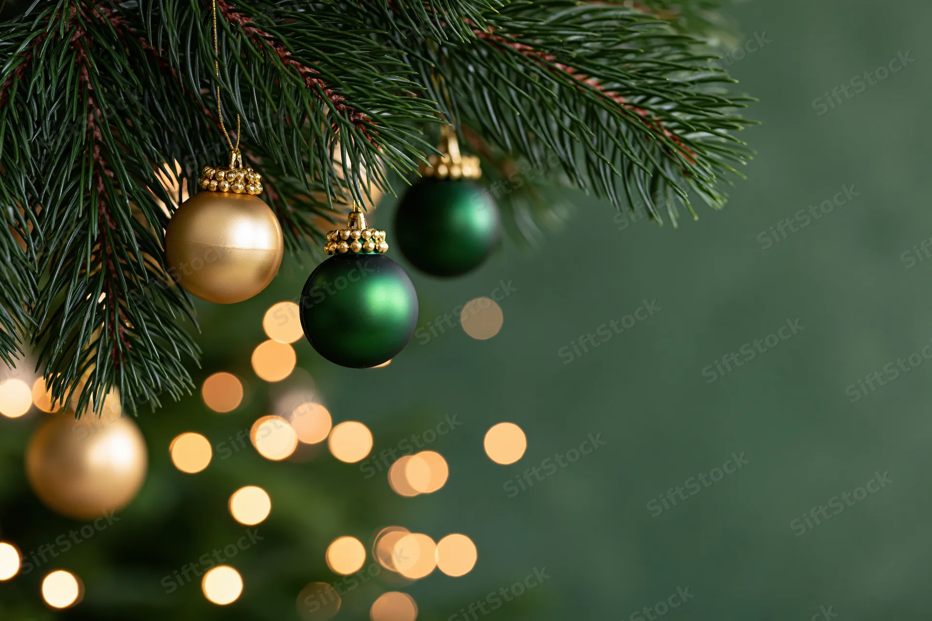 Pine tree branches with gold and green ornaments hanging against blurred warm lights and green background